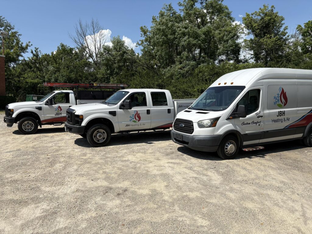Three white JBH Heating & Air service vans parked in a gravel lot with trees and blue sky in the background.