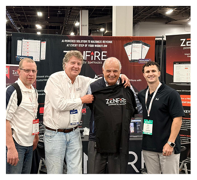 Group of four men at a tech expo booth posing for a photo; the second from left and the center man hold a black ZenFire shirt, with booth banners and device screens visible in the background.