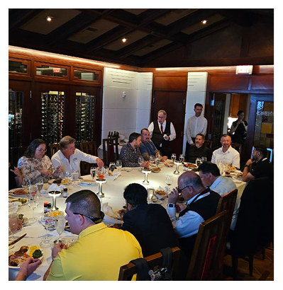 Group of people seated around a round table in a formal dining room, enjoying wine and a shared meal as servers stand nearby.