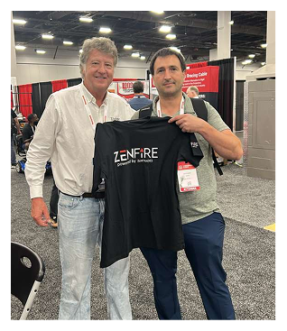 Two men at a convention posing together while holding up a black Zenfire t-shirt and smiling, with badges and exhibitor booths in the background.