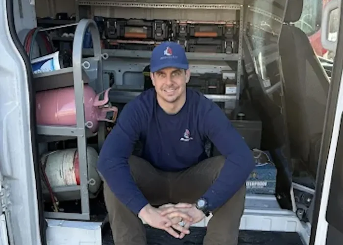 Man in a blue cap and navy long-sleeve shirt sits cross-legged in the open back of a work van, with shelves of tools and equipment behind him.