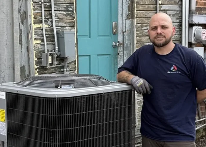 HVAC technician in a dark shirt and gloves stands next to a large outdoor air conditioning condenser near a weathered brick wall and teal door.