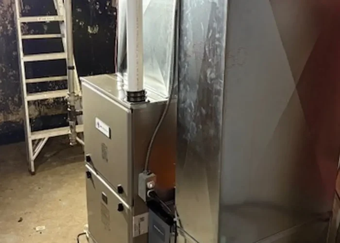 Industrial furnace with metal ductwork in a utility room; a white ladder sits against a dark wall in the background.
