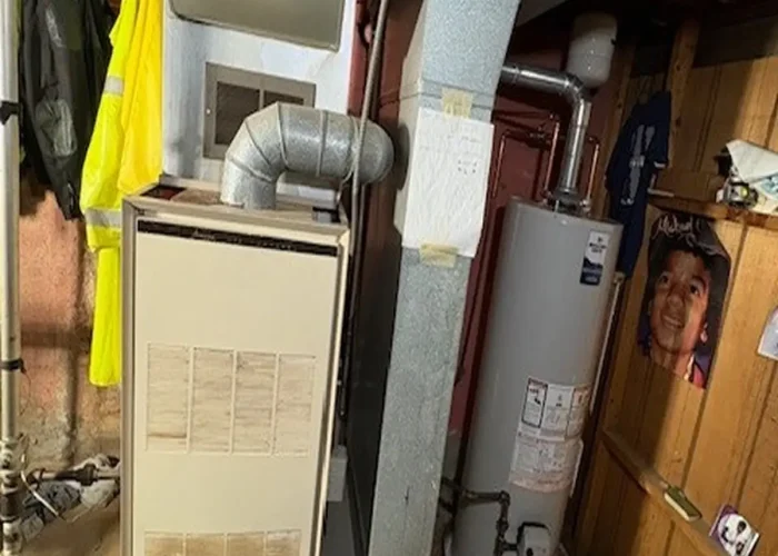 Basement utility area showing a beige furnace with ductwork, a water heater nearby, and a yellow safety vest hanging on the wall.