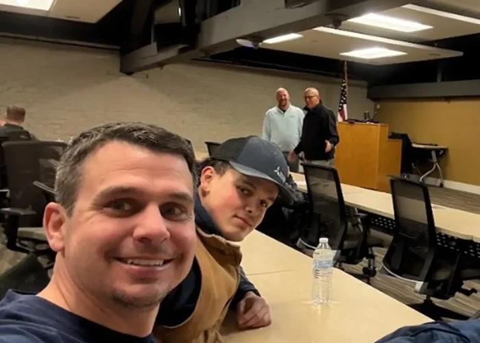 Man in the foreground takes a selfie at a long conference table as two men stand near a podium with an American flag in the background.